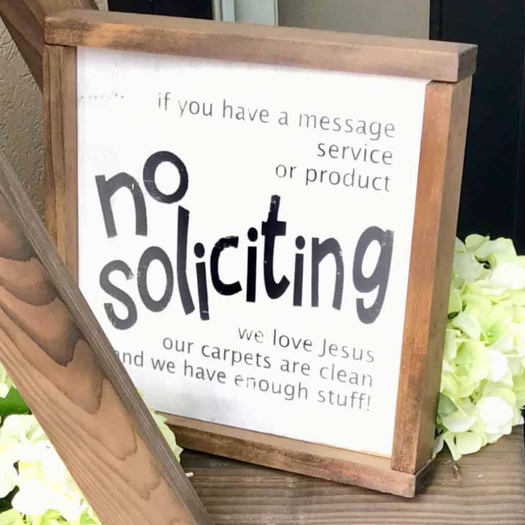 No Soliciting sign made out of wood and sitting on a shelf with flowers nearby.