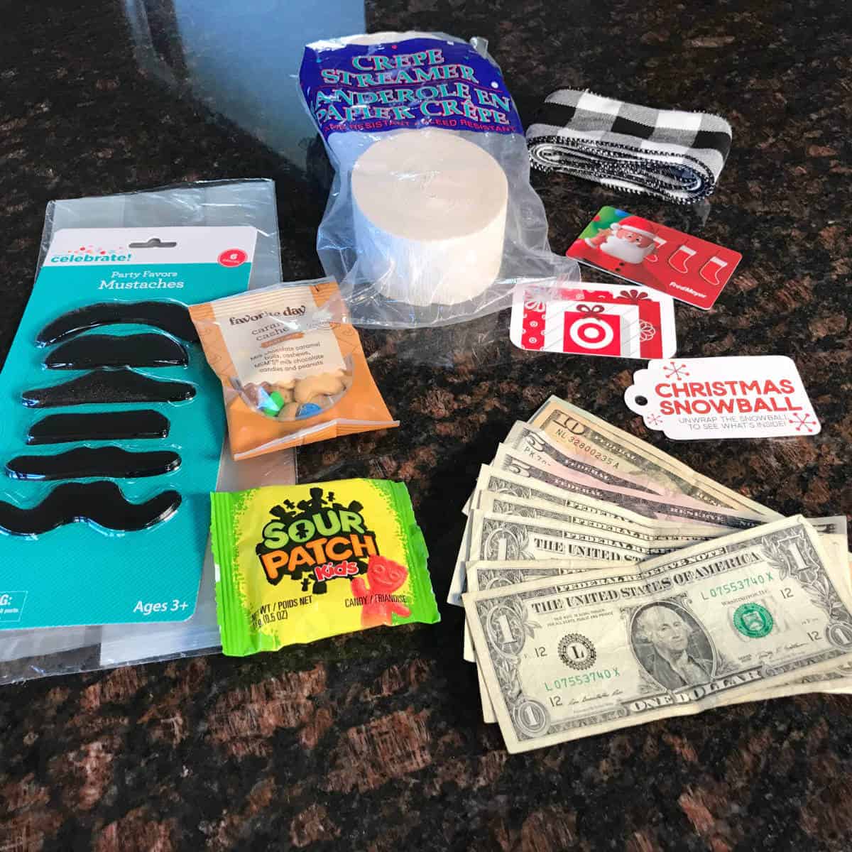 A package of stick on mustaches, white paper streamer, money, Target gift card, candy, trail mix, and a Christmas Snowball tag lying on a counter.