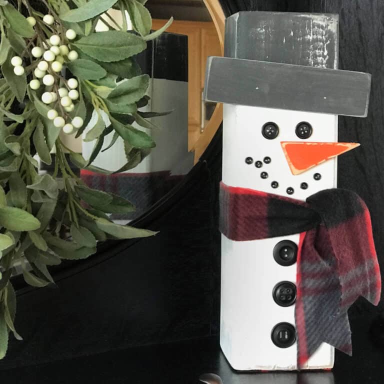 Picture of a snowman, made out of a piece of 4x4 wood, sitting in front of a mirror next to a green wreath.