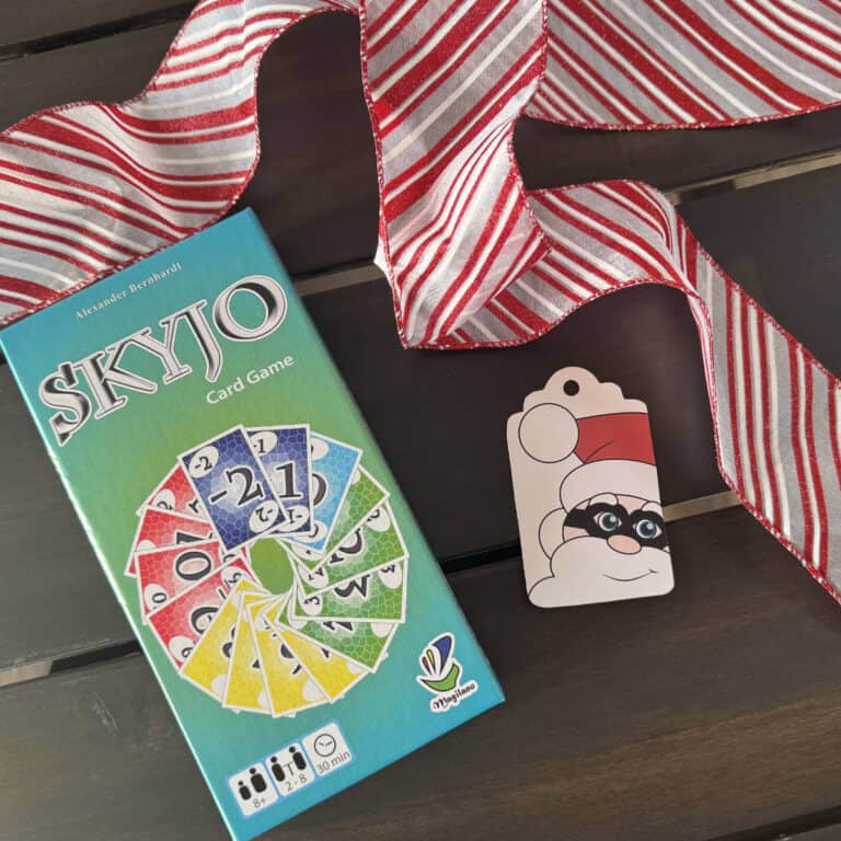 SKYJO game sitting on a table next to a printed gift tag with Santa wearing a thief mask and red and white ribbon.