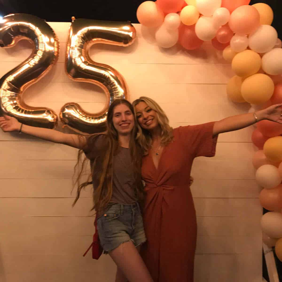 Two girls standing in front of a wood backdrop wall that's been decorated with balloons.