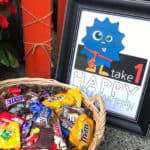 Printable sign that's been framed and says Take 1, Happy Halloween by a basket of treats.