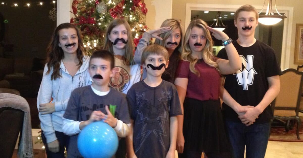 Group of kids and teenagers wearing fake mustaches in front of a Christmas tree.