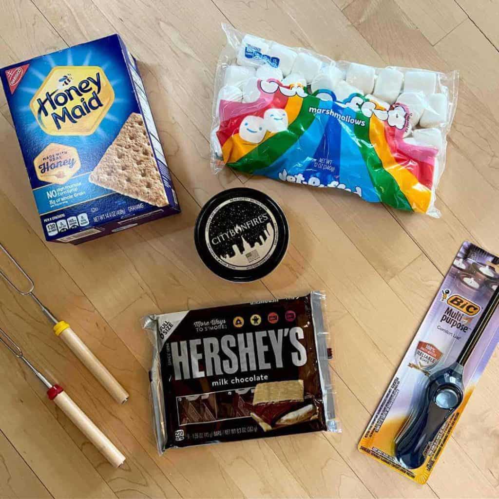 S'more care package supplies: graham crackers, marshmallows, lighter, roasting sticks, chocolate, and personal fire pit.