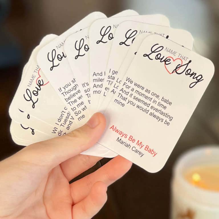 Name That Love Song game cards in someone's hand with a lit candle in the background.