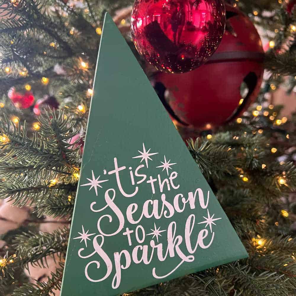 Green triangle wood Christmas tree with the words 'tis the season to sparkle' painted on it.