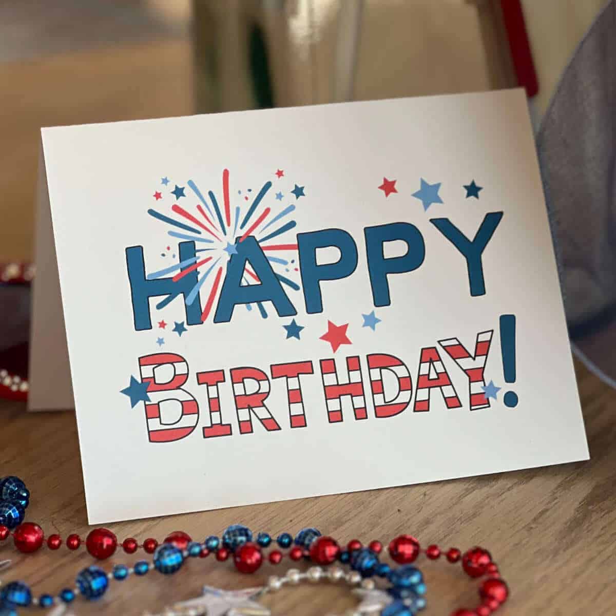 Printable Happy Birthday Card for the 4th of July sitting on a table next to red, white, and blue necklaces.