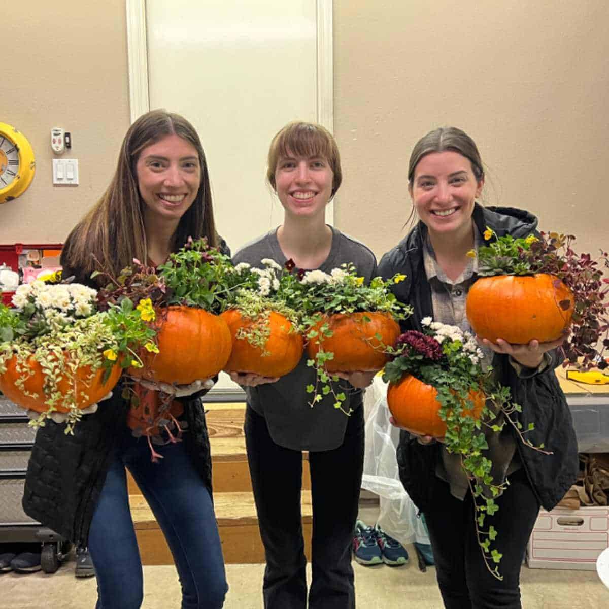 Three young ladies holding pumpkins with plants planted inside.