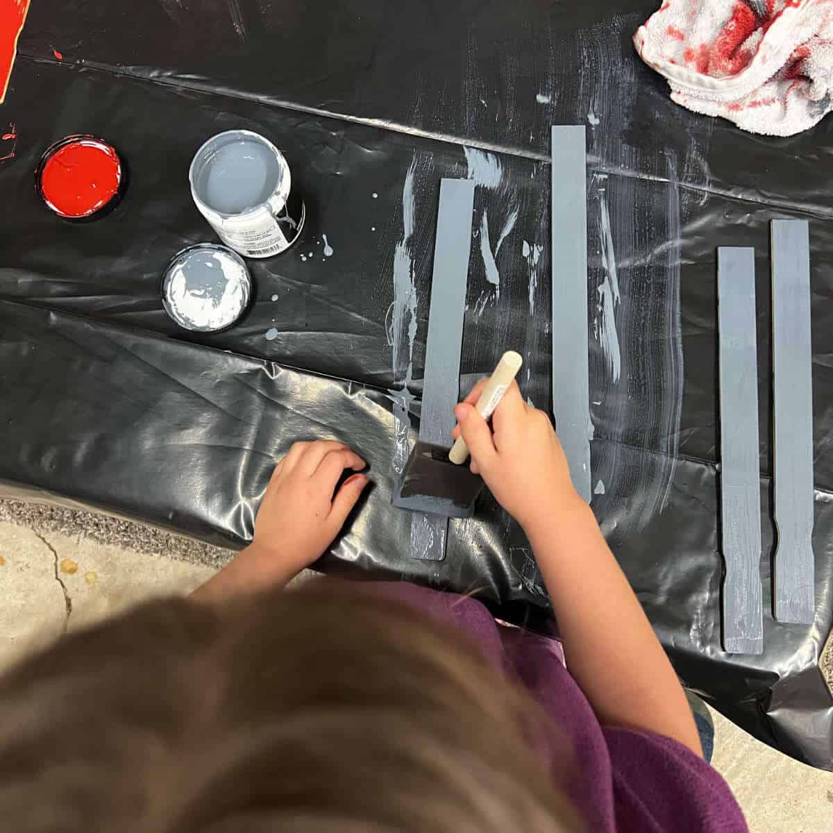 Child painting paint sticks blue using a sponge paintbrush.