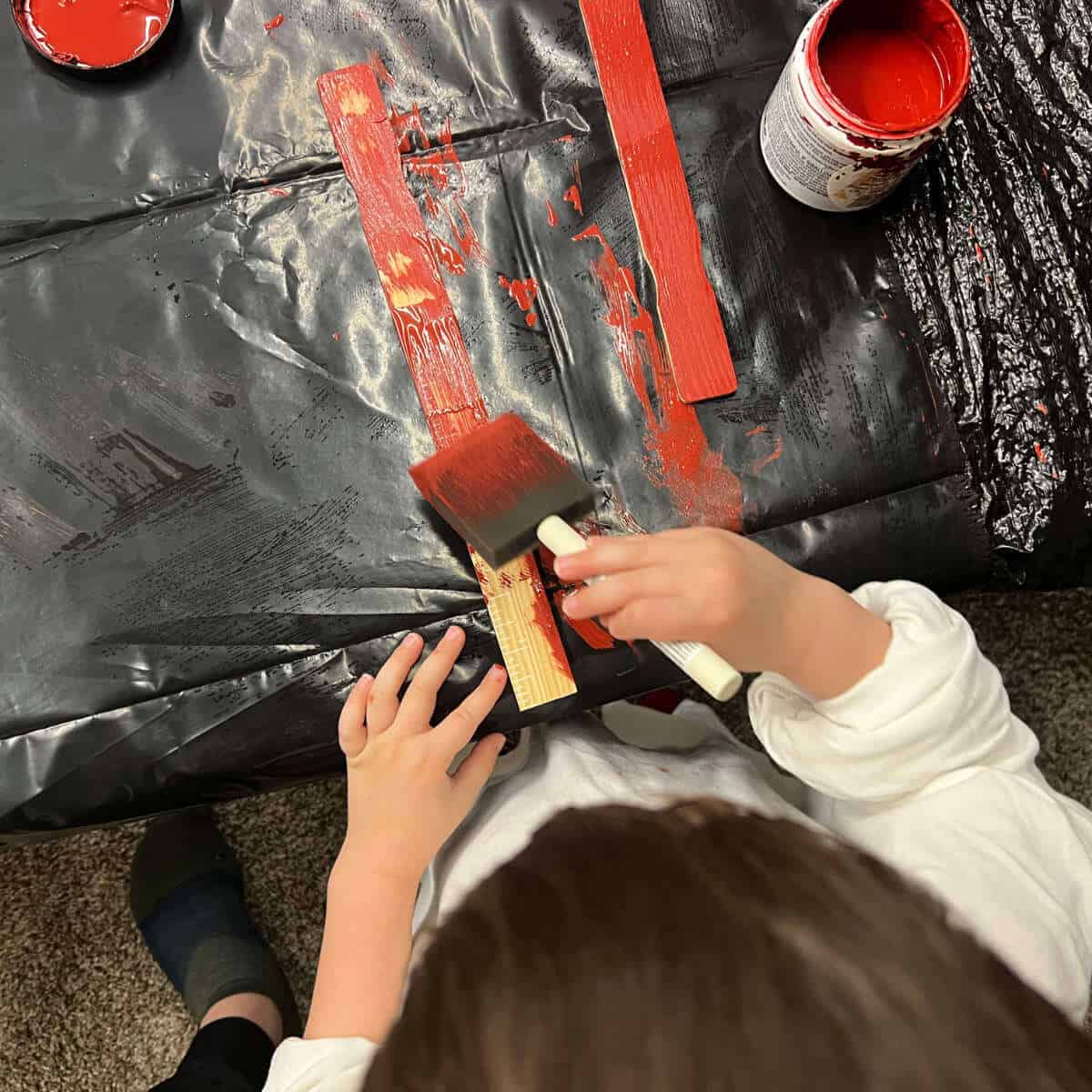 Child painting two paint sticks red using a sponge paintbrush.