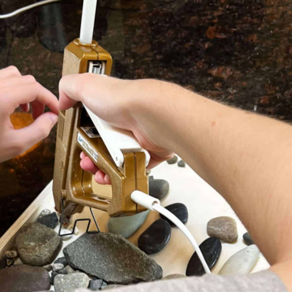 Girl's hands holding a glue gun to glue rocks to a wood canvas.