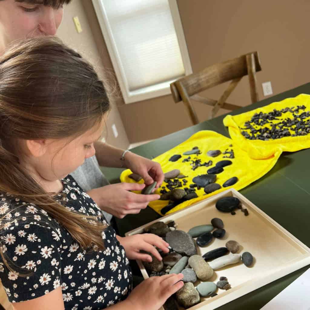 Little girl and her nanny arranging rocks in a wood canvas to create a family portrait.