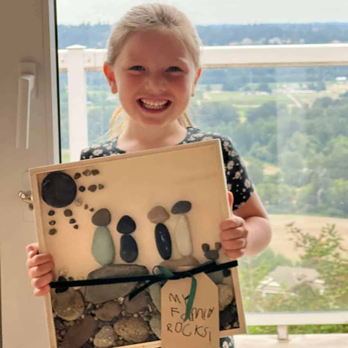 Little girl holding a wood canvas with a rock family portrait glued on.