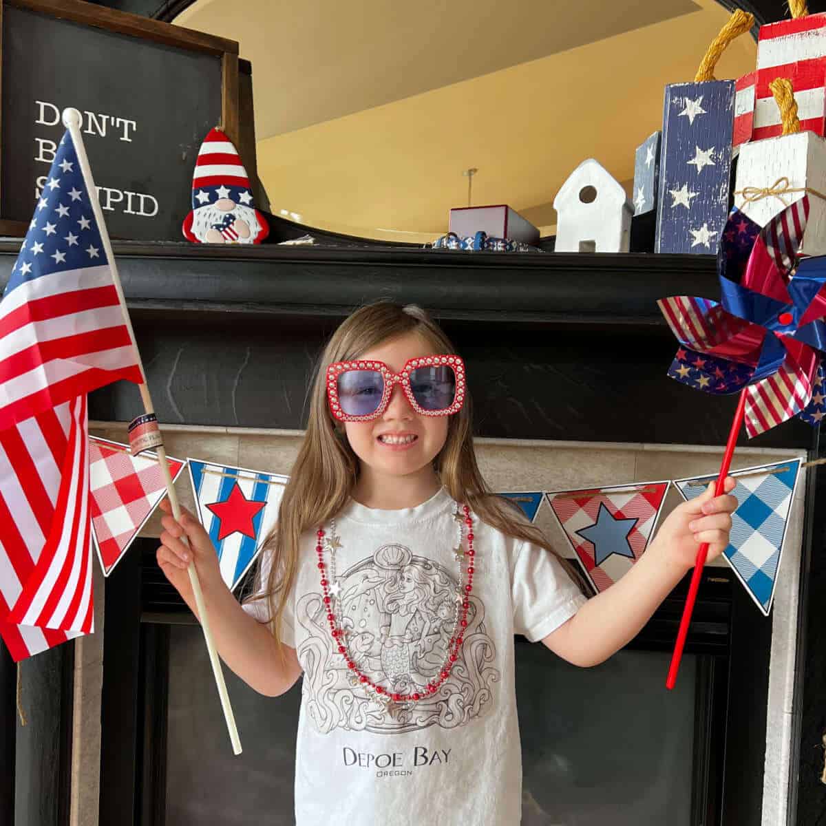 Little girl holding an American flag and pinwheel while wearing a festive necklace and sunglasses in preparation for the 4th of July.