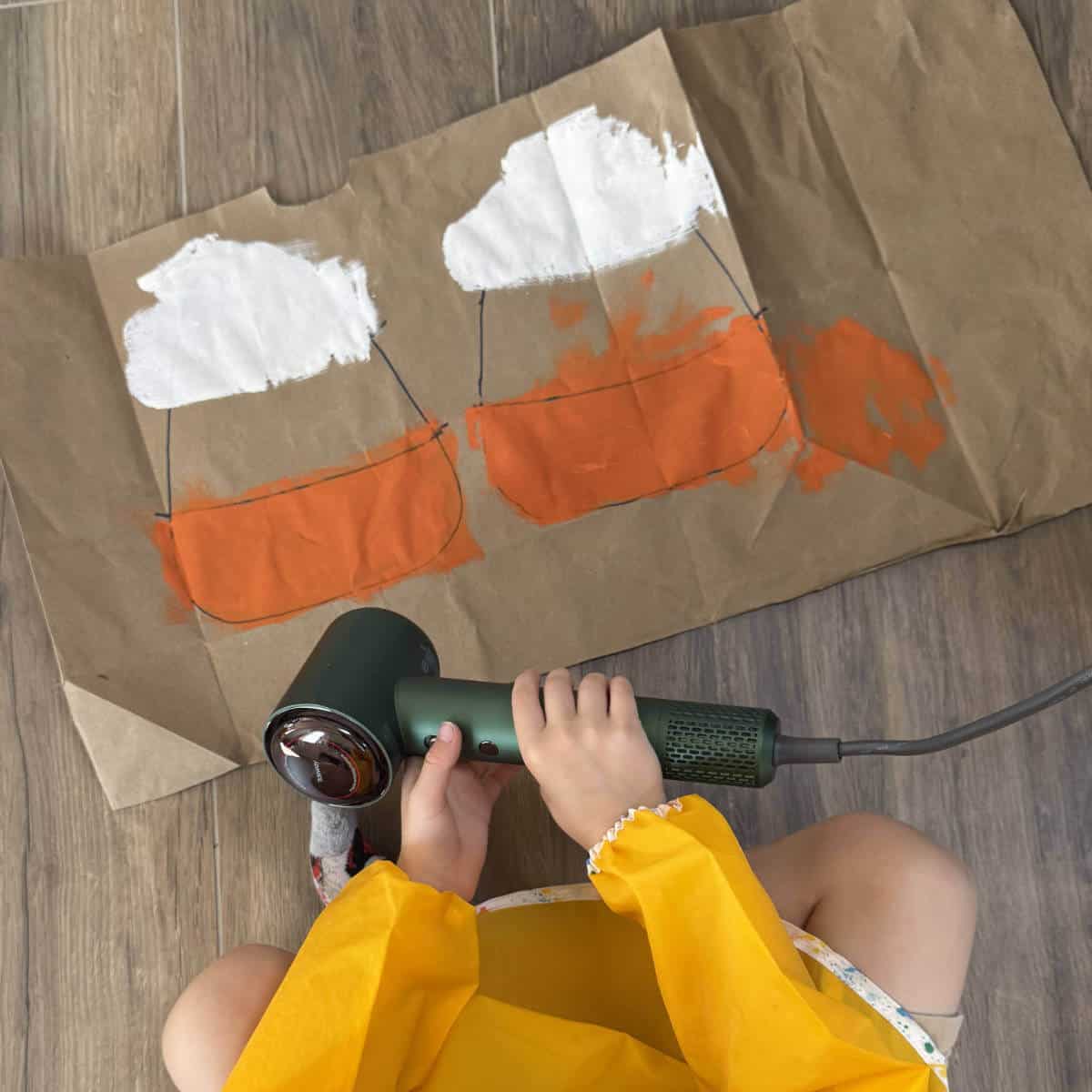 A child using a hairdryer to dry the paint on a brown paper bag.