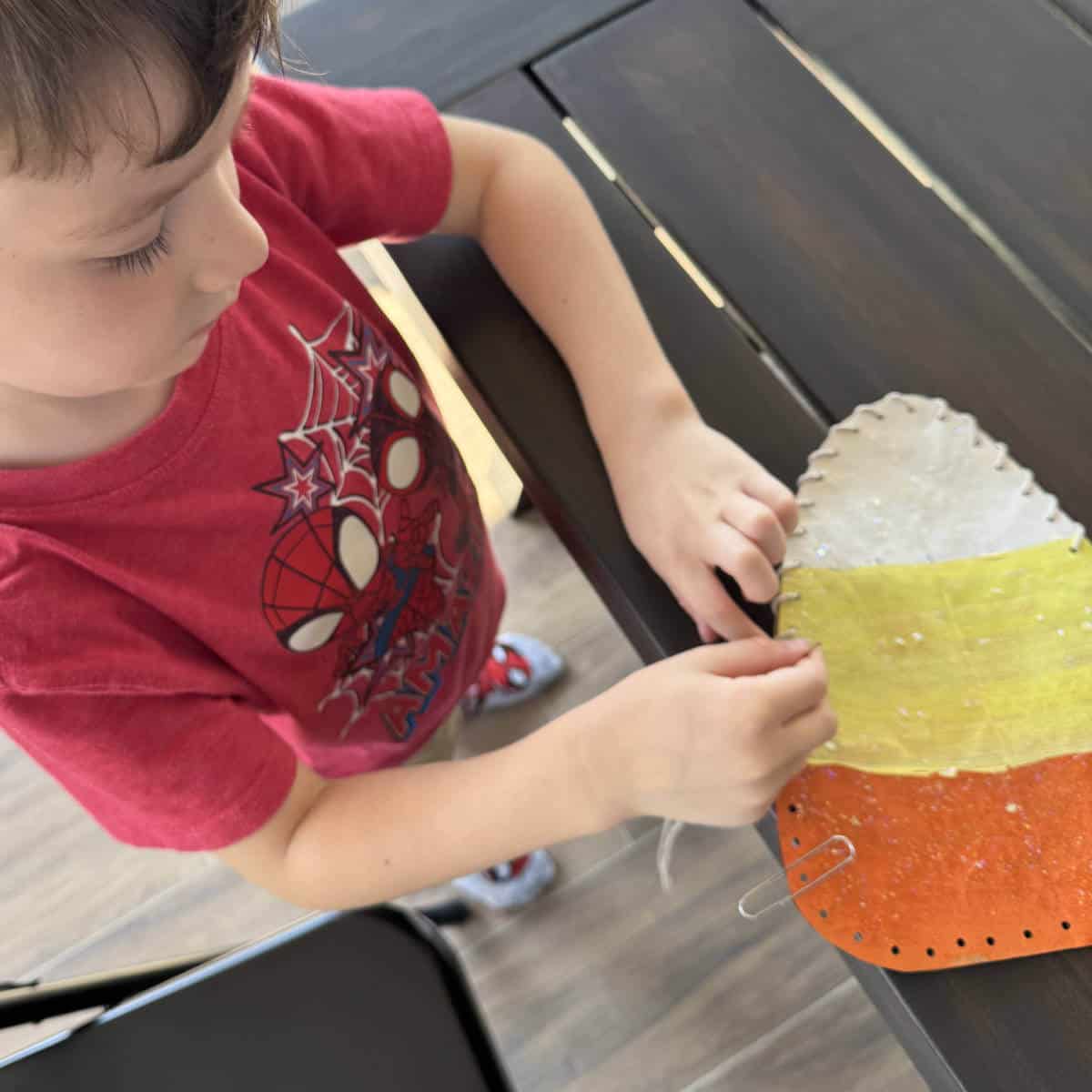 Boy sewing together the edges of a paper bag candy corn craft using yarn.