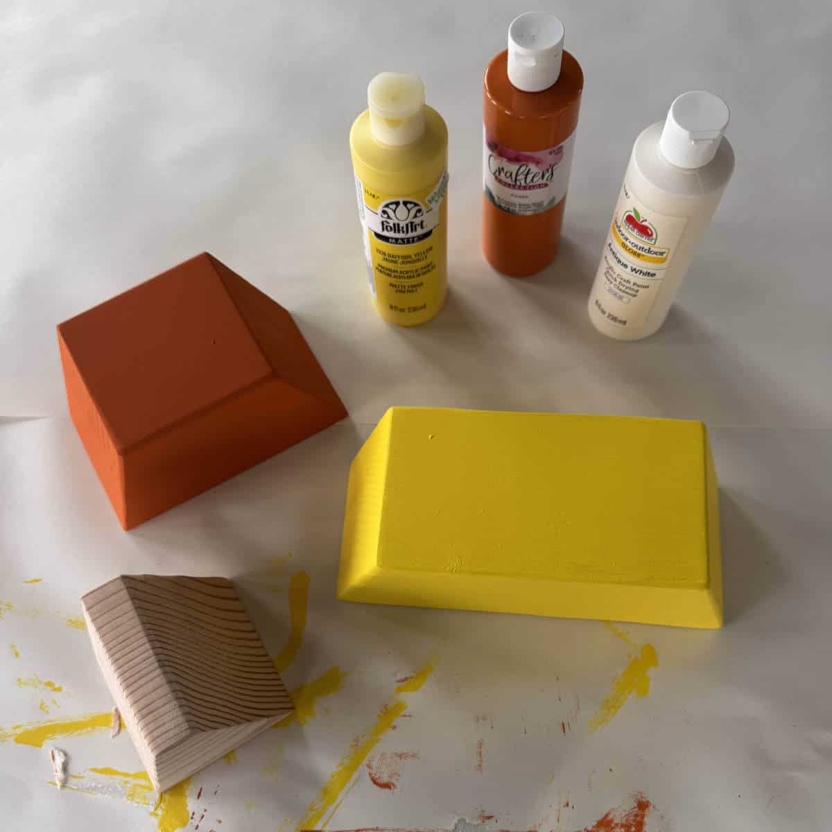 Image of three pieces of a 4x4 candy corn with the bottom piece painted yellow, the middle piece painted orange, and the top piece ready to be painted. There are three bottles of acrylic paint nearby.