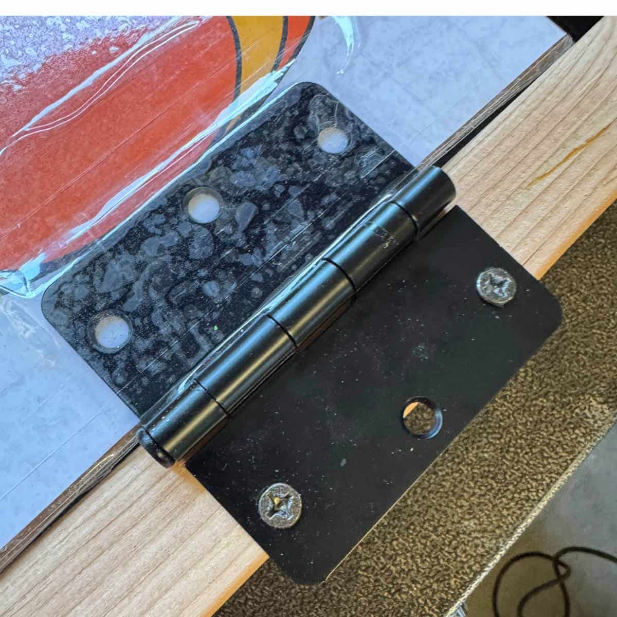 Image of a door hinge attached to a paper flame with packing tape on one side and screwed to a 2x4 on the other side.