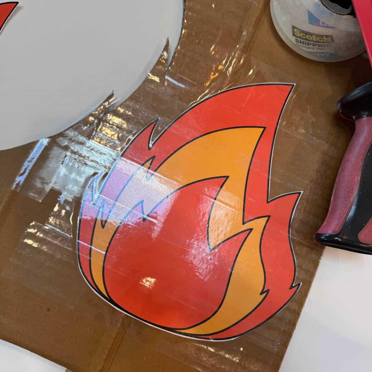 Image of a clipart flame that has been cut out and taped to cardboard using packing tape.